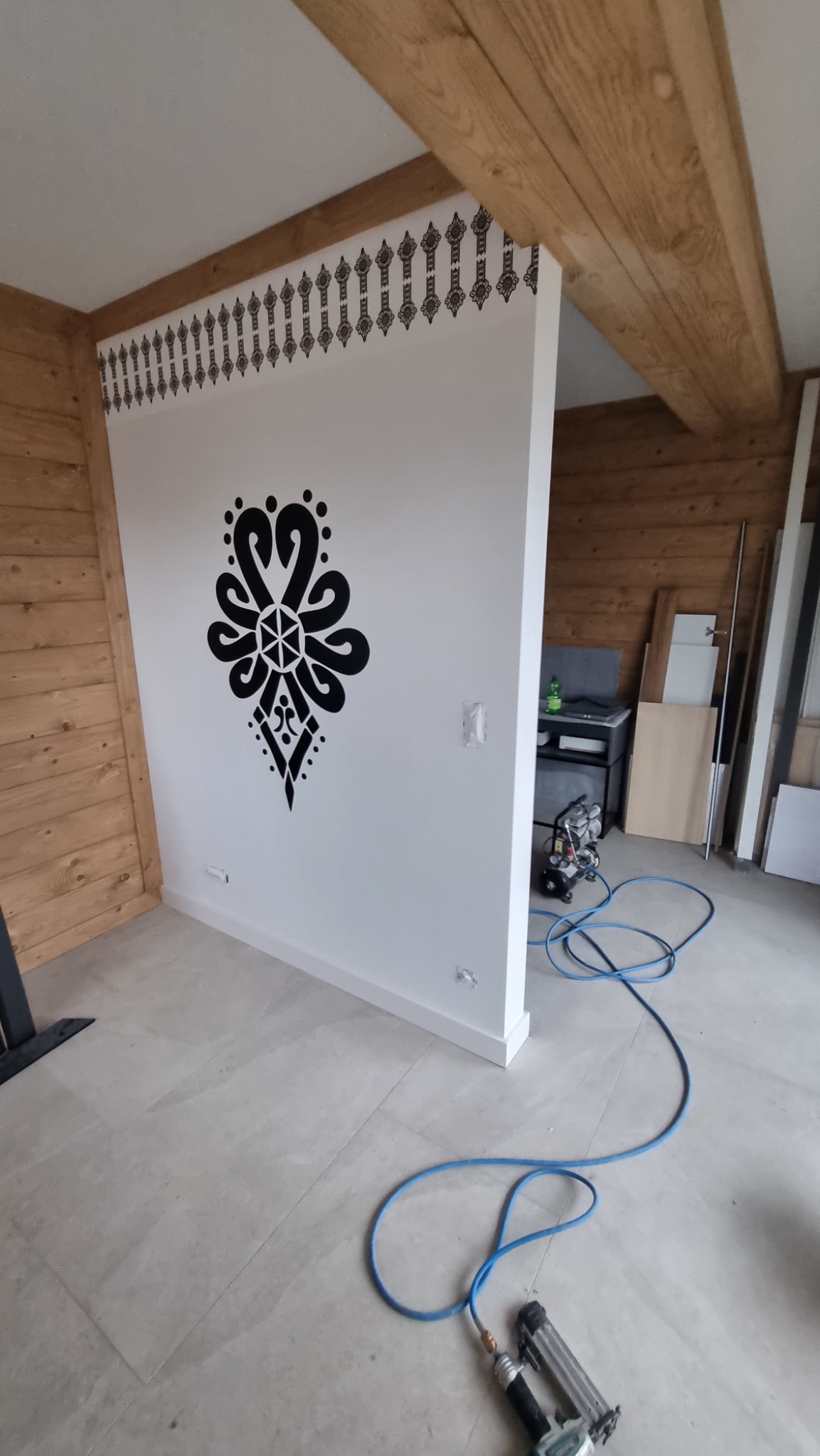 Ściana działowa w rustykalnym wnętrzu z folkowym motywem dekoracyjnym, widoczny kompresor i pistolet pneumatyczny do gwoździ.