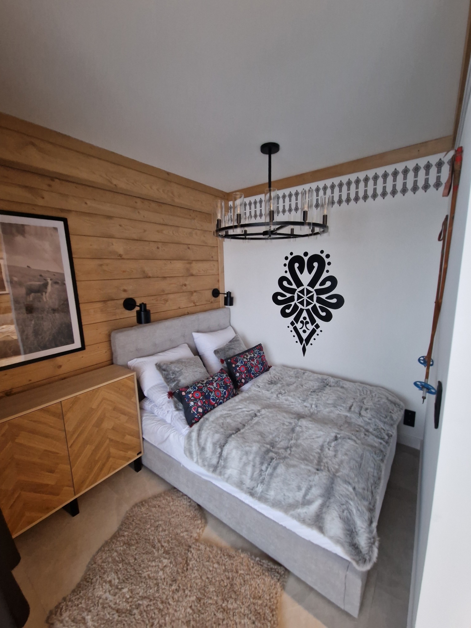 Sypialnia z drewnianą ścianą, komodą w jodełkę, łóżkiem przykrytym futrzaną narzutą i dekoracyjnym wzorem góralskim na ścianie.