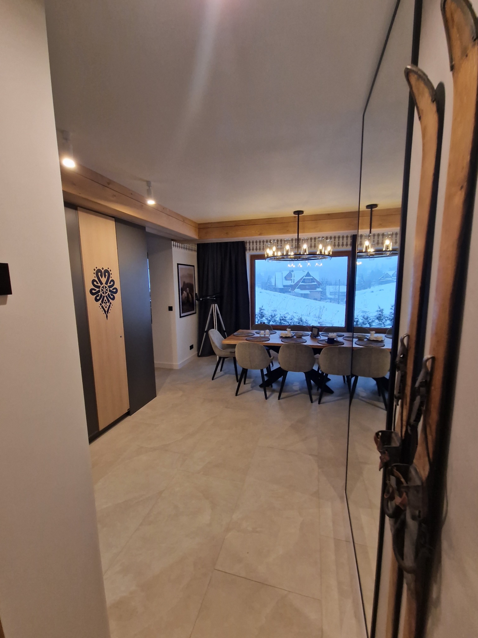Nowoczesna jadalnia z widokiem na ośnieżony krajobraz górski, parkiet w odcieniach beżu, dekoracyjne narty przy ścianie, przesuwne drzwi z folkowym motywem.