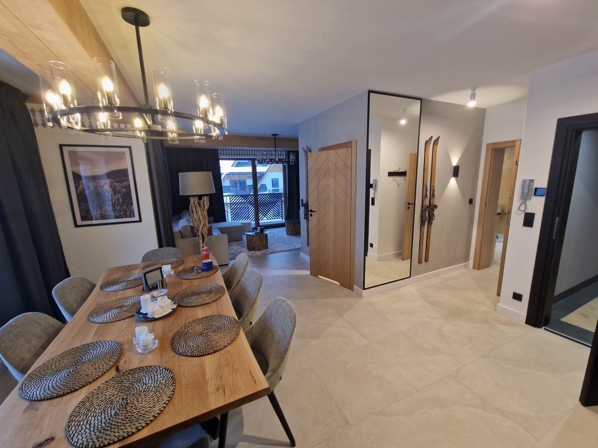 Jasne, przestronne wnętrze apartamentu w Nowym Targu z drewnianym stołem jadalnym, dekoracyjnym oświetleniem i widokiem na zaśnieżone góry przez balkon.