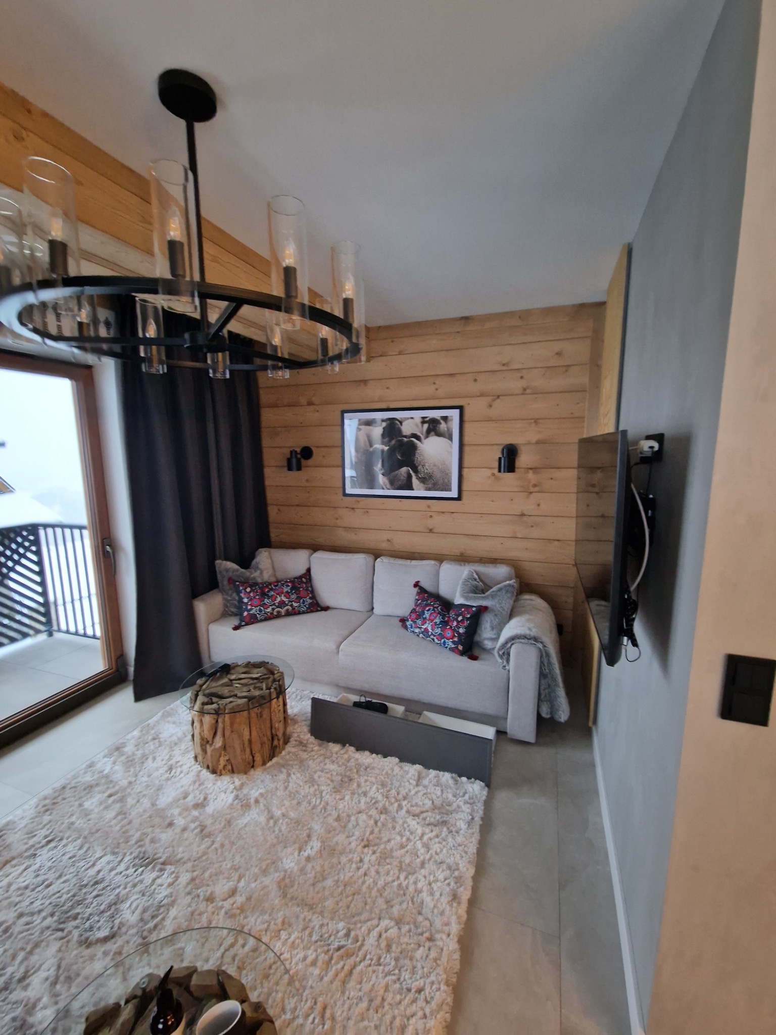 Jasny salon z drewnianą ścianą, szarą sofą z dekoracyjnymi poduszkami, puszystym dywanem i stolikiem kawowym imitującym pień drzewa, oświetlony nowoczesnym żyrandolem.