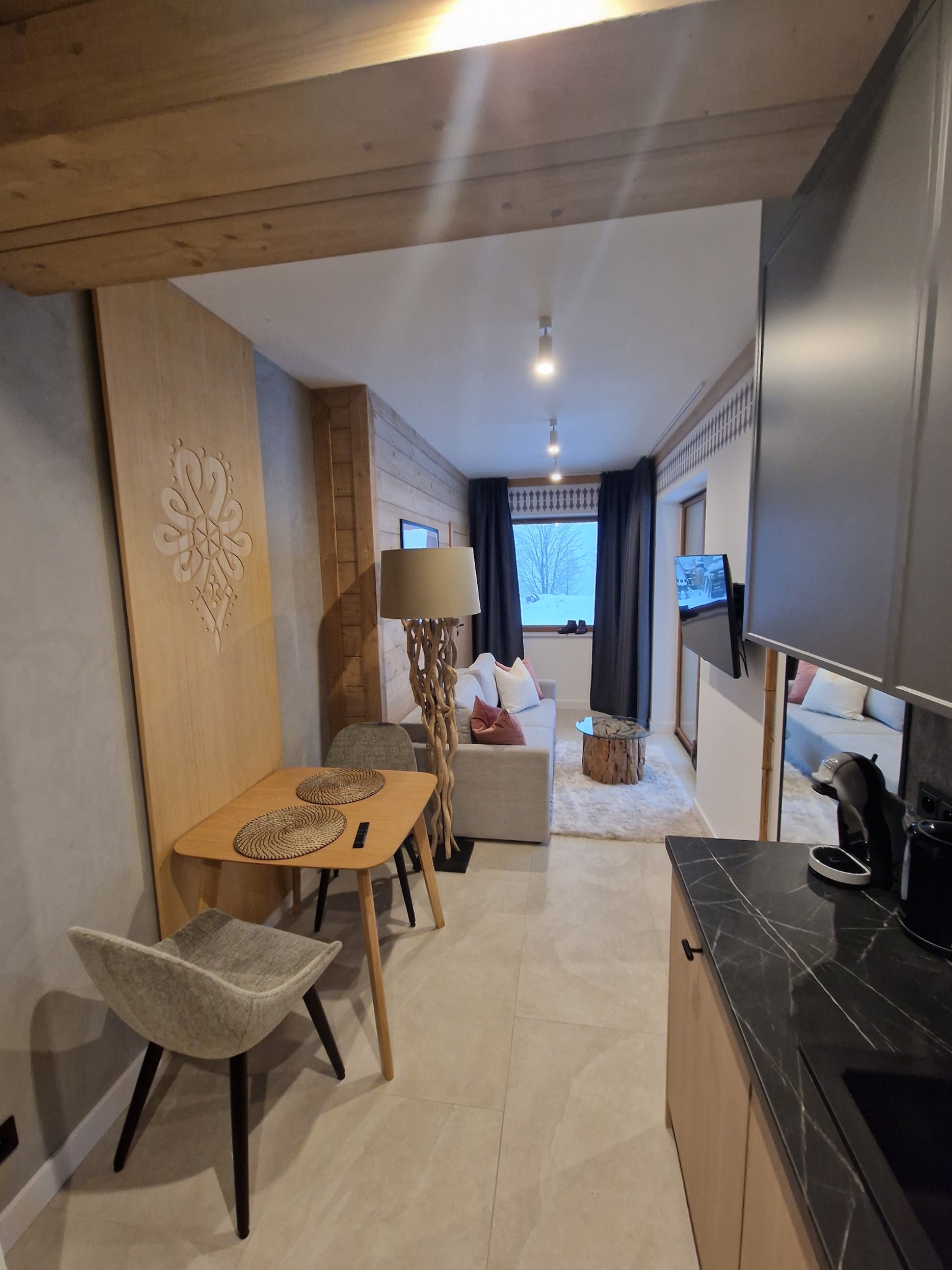 Nowoczesny salon z drewnianymi elementami dekoracyjnymi, w tle widok na ośnieżony krajobraz za oknem, stolik z plecionymi podkładkami i lampa z podstawą z gałęzi.