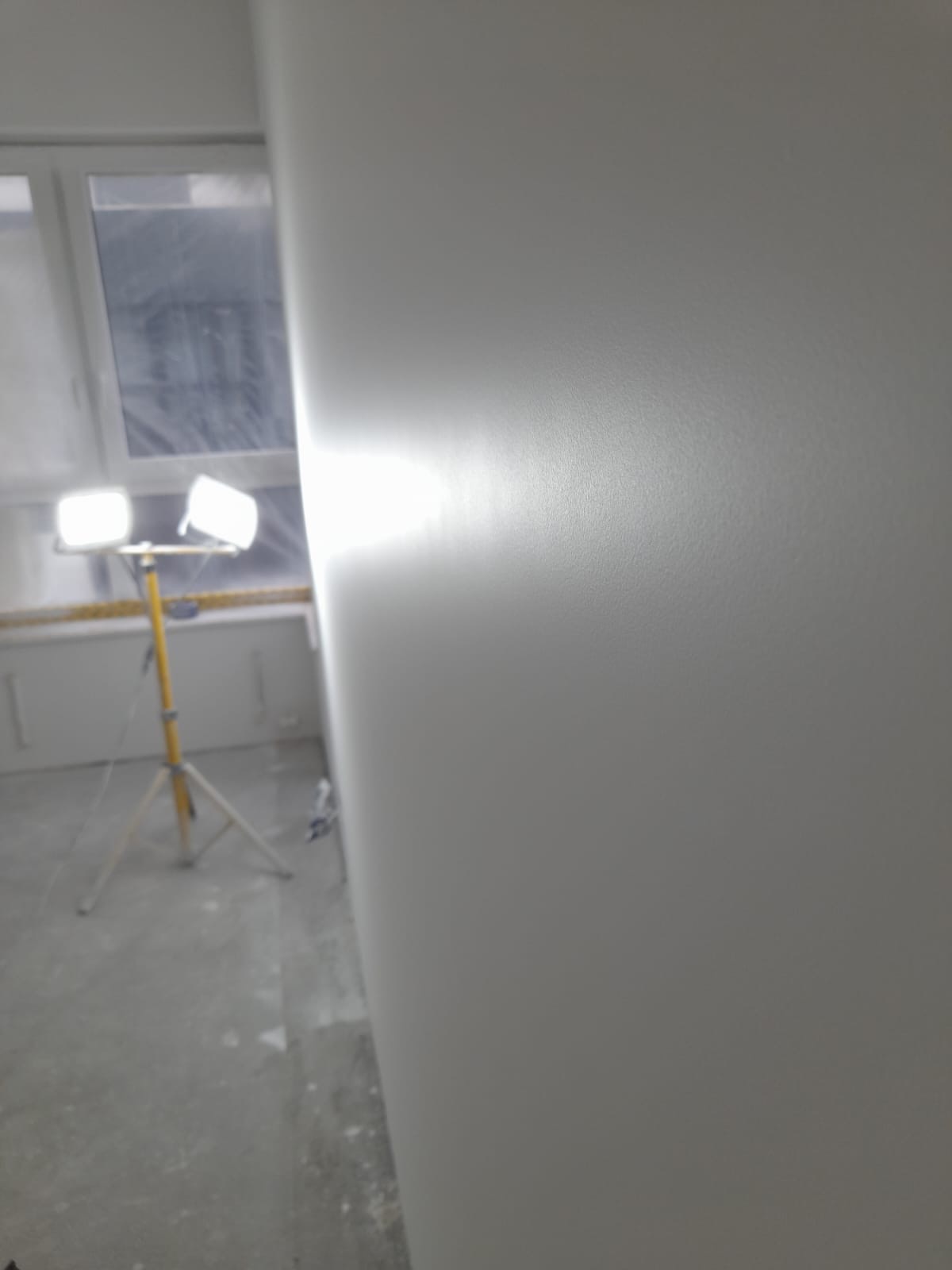 Świeżo pomalowana, gładka ściana w odcieniach szarości, oświetlona przez lampę budowlaną na statywie w tle, z widocznym oknem zabezpieczonym folią.