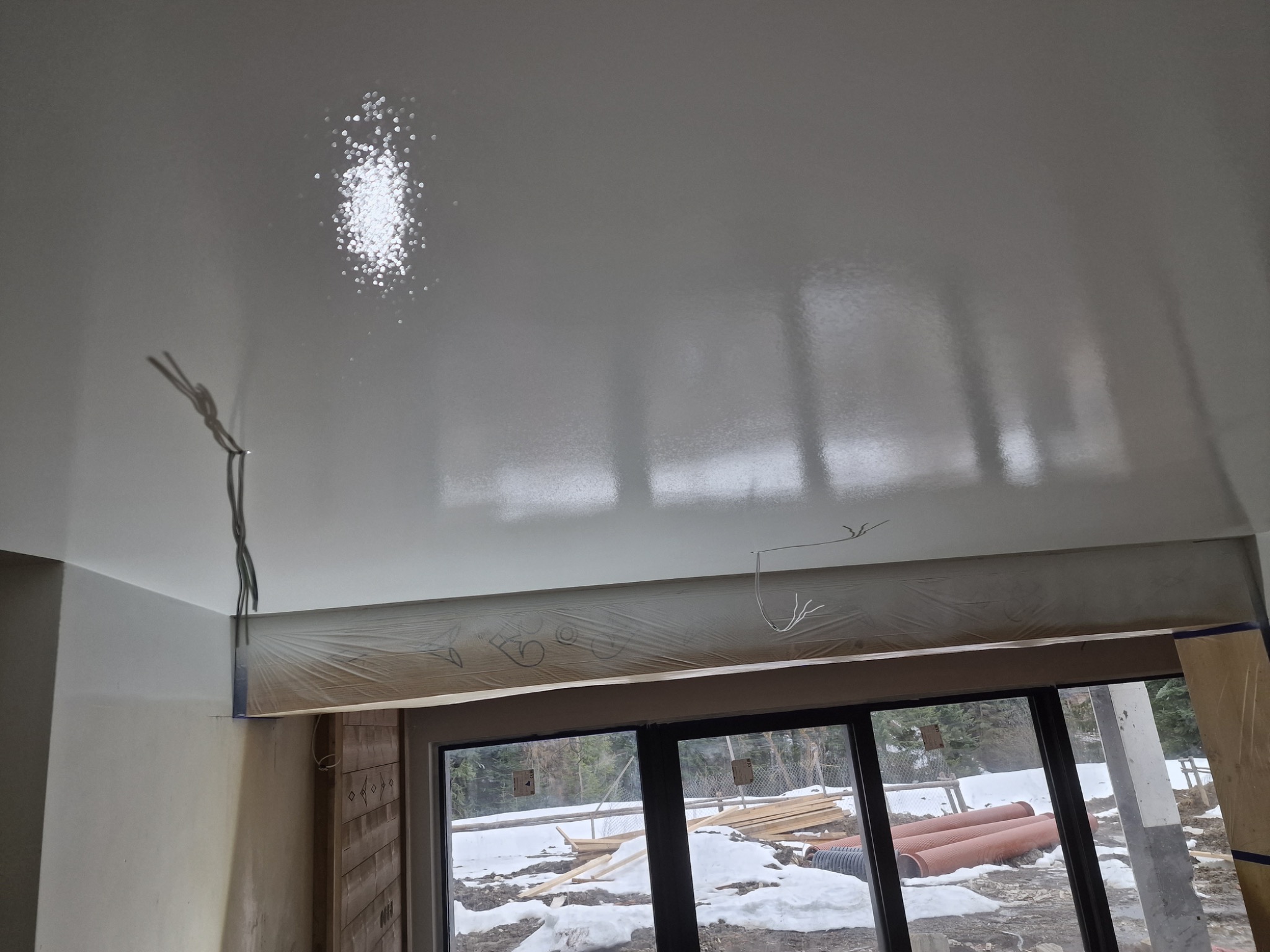 Świeżo pomalowany, błyszczący sufit z widocznymi kablami, zabezpieczonymi folią framugami okiennymi i zimowym krajobrazem za oknem.