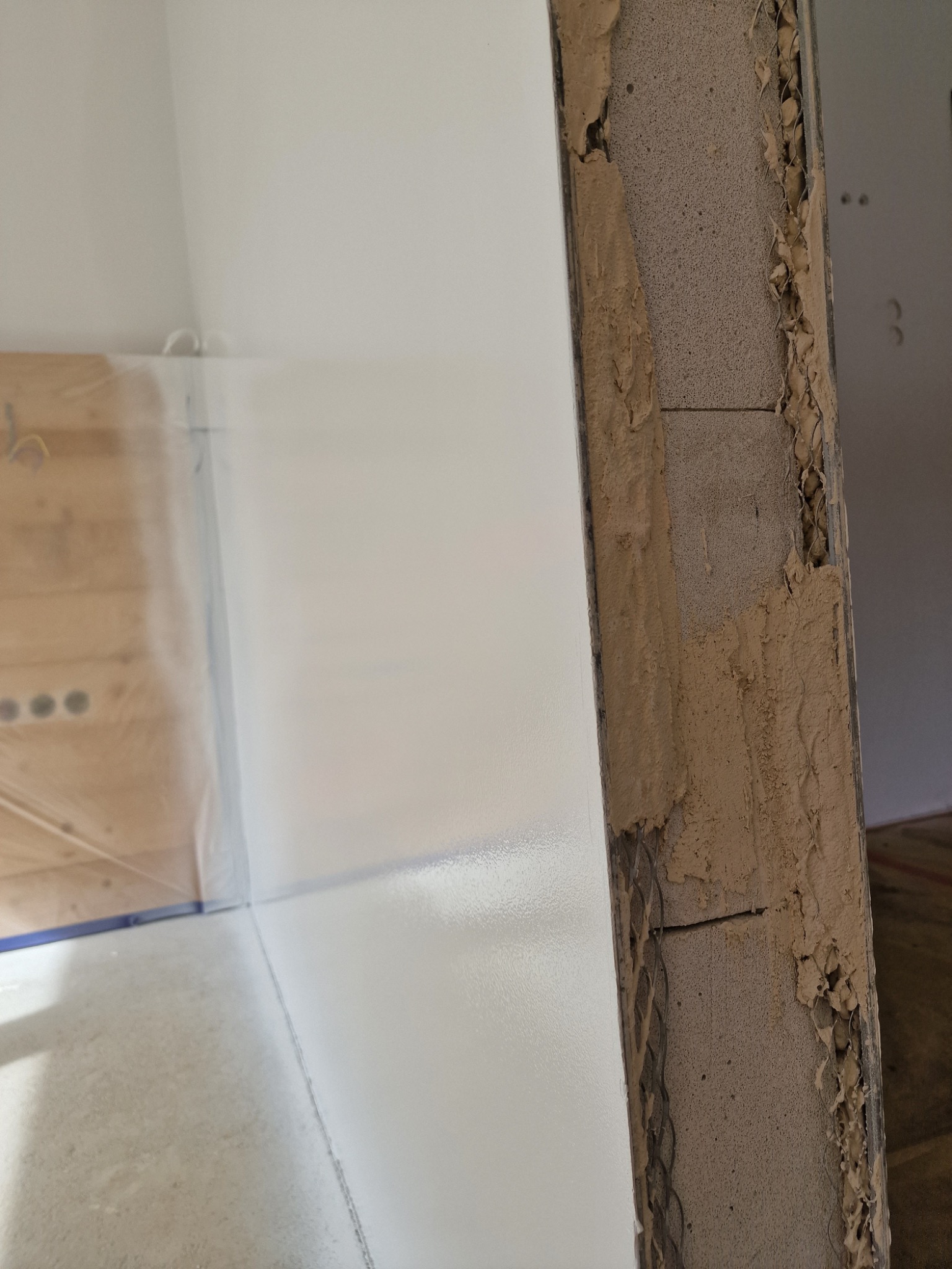 Świeżo pomalowana, biała ściana z widocznym narożnikiem z surowego betonu i resztkami zaprawy, zabezpieczona folią podłoga.