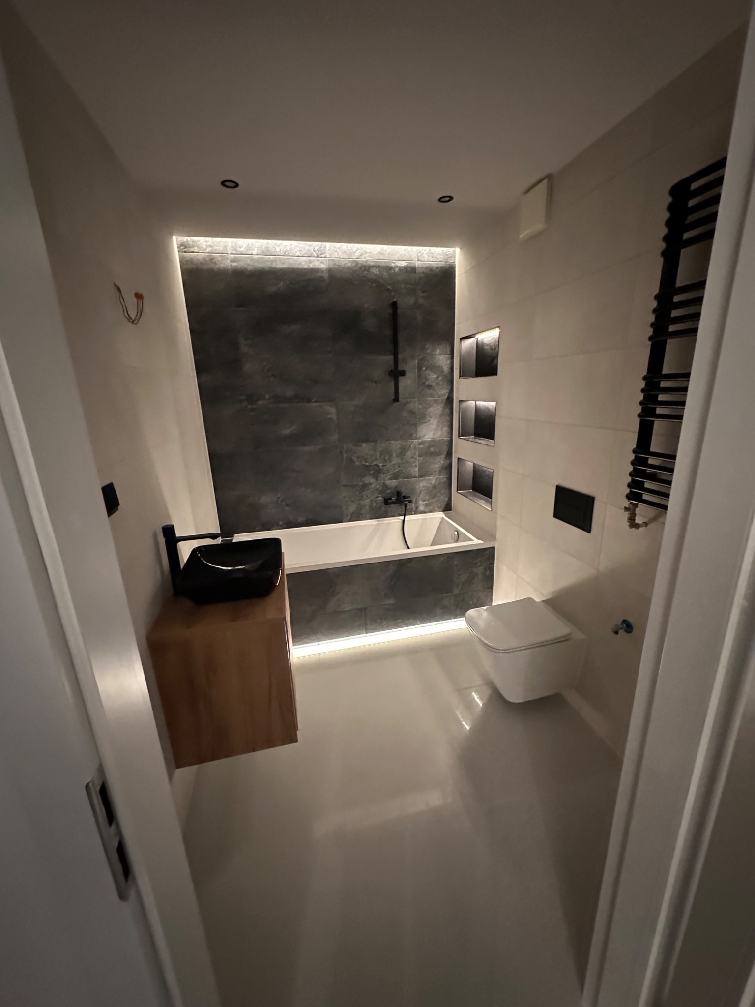 Nowoczesna łazienka z podświetlaną wanną, czarną armaturą i umywalką nablatową na drewnianej szafce, widok zza uchylonych drzwi.