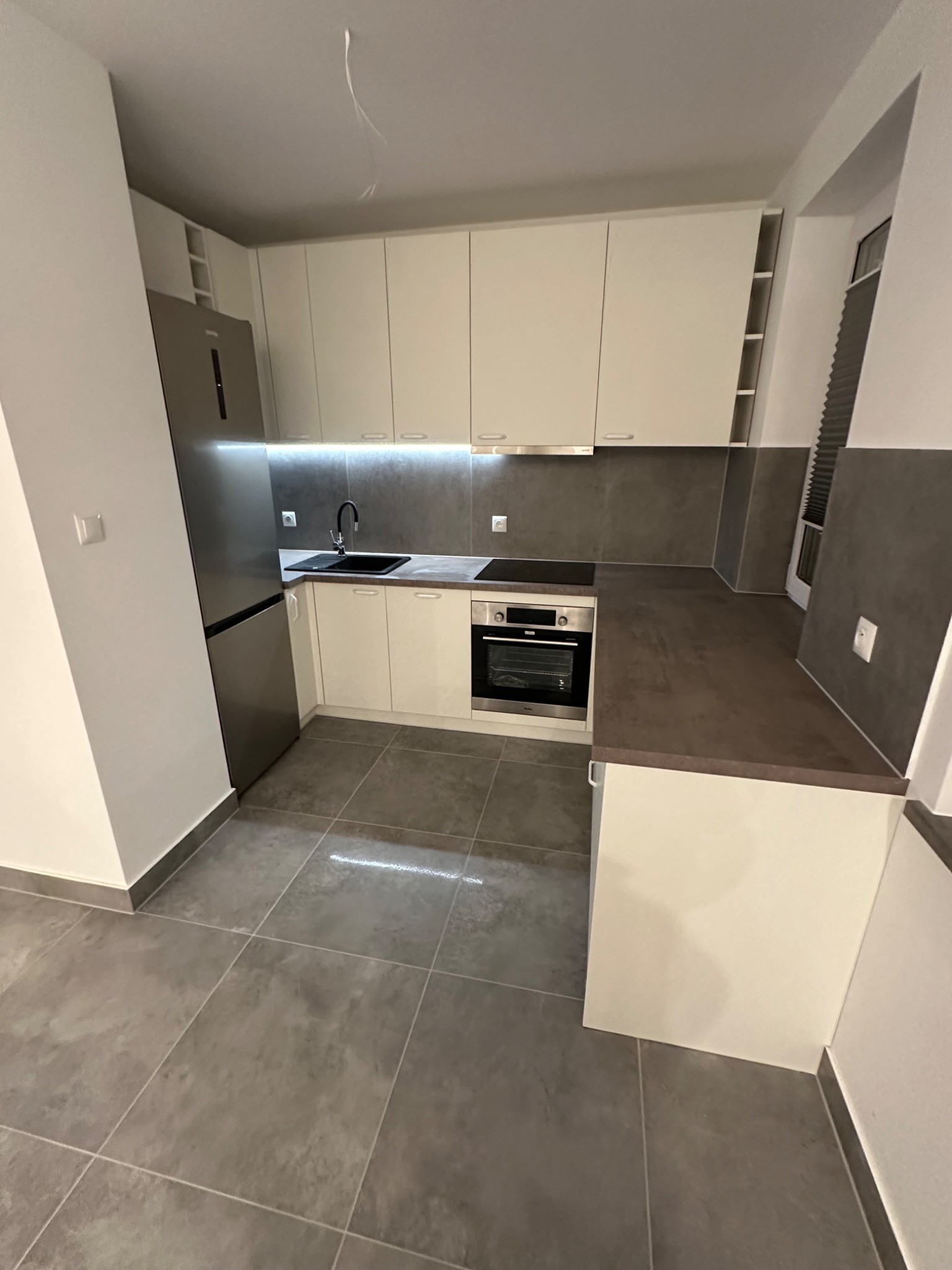 Nowoczesna, jasna kuchnia z szarymi elementami, obejmująca zabudowę z białymi szafkami, szarym blatem, zintegrowanym oświetleniem LED, stalową lodówką i szarą podłogą z płytek.