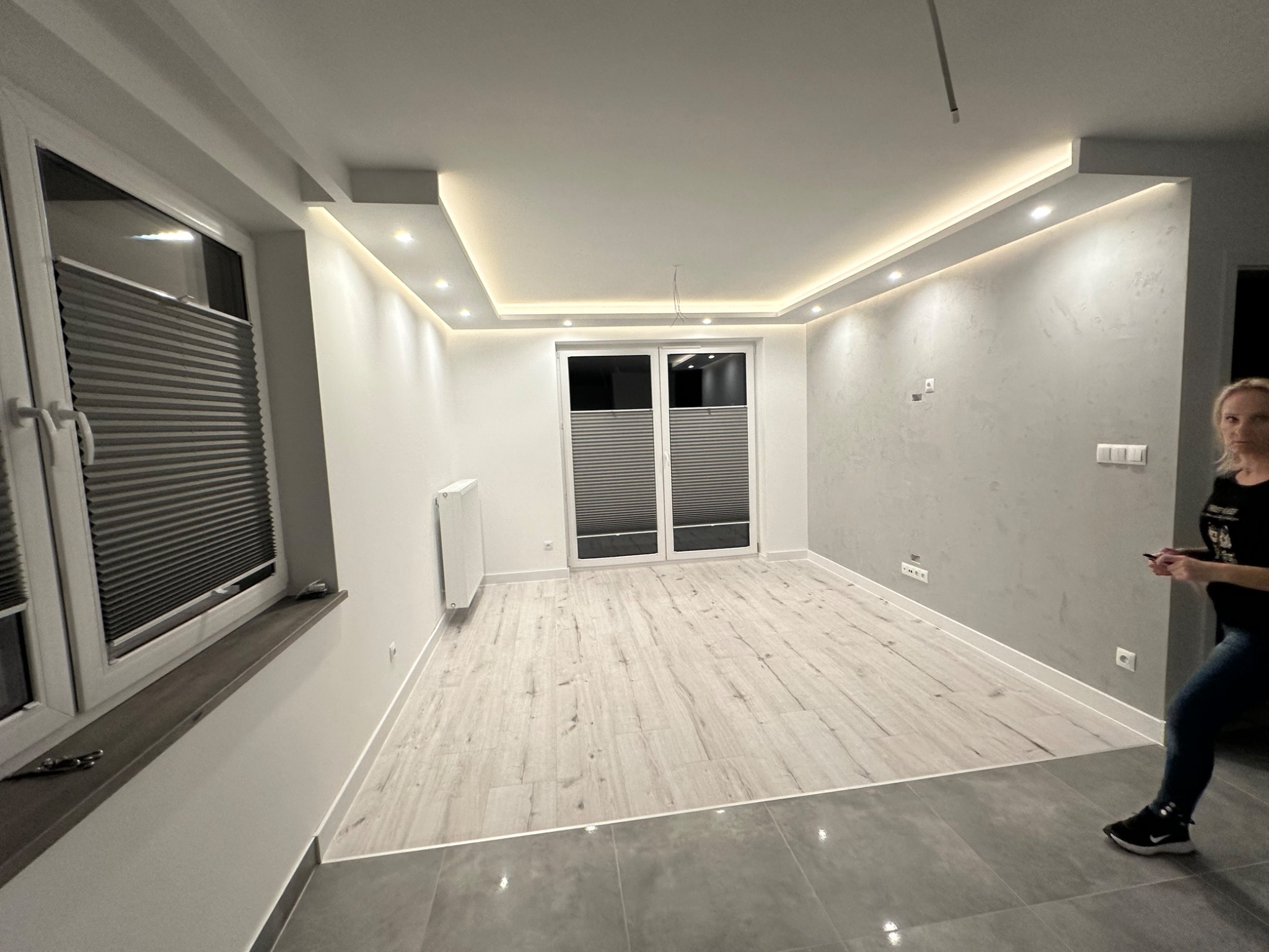Jasne, przestronne wnętrze po remoncie: parkiet imitujący drewno, szara ściana z fakturą, podwieszany sufit z oświetleniem LED, okna z plisowanymi roletami, szare płytki podłogowe przy wejściu.