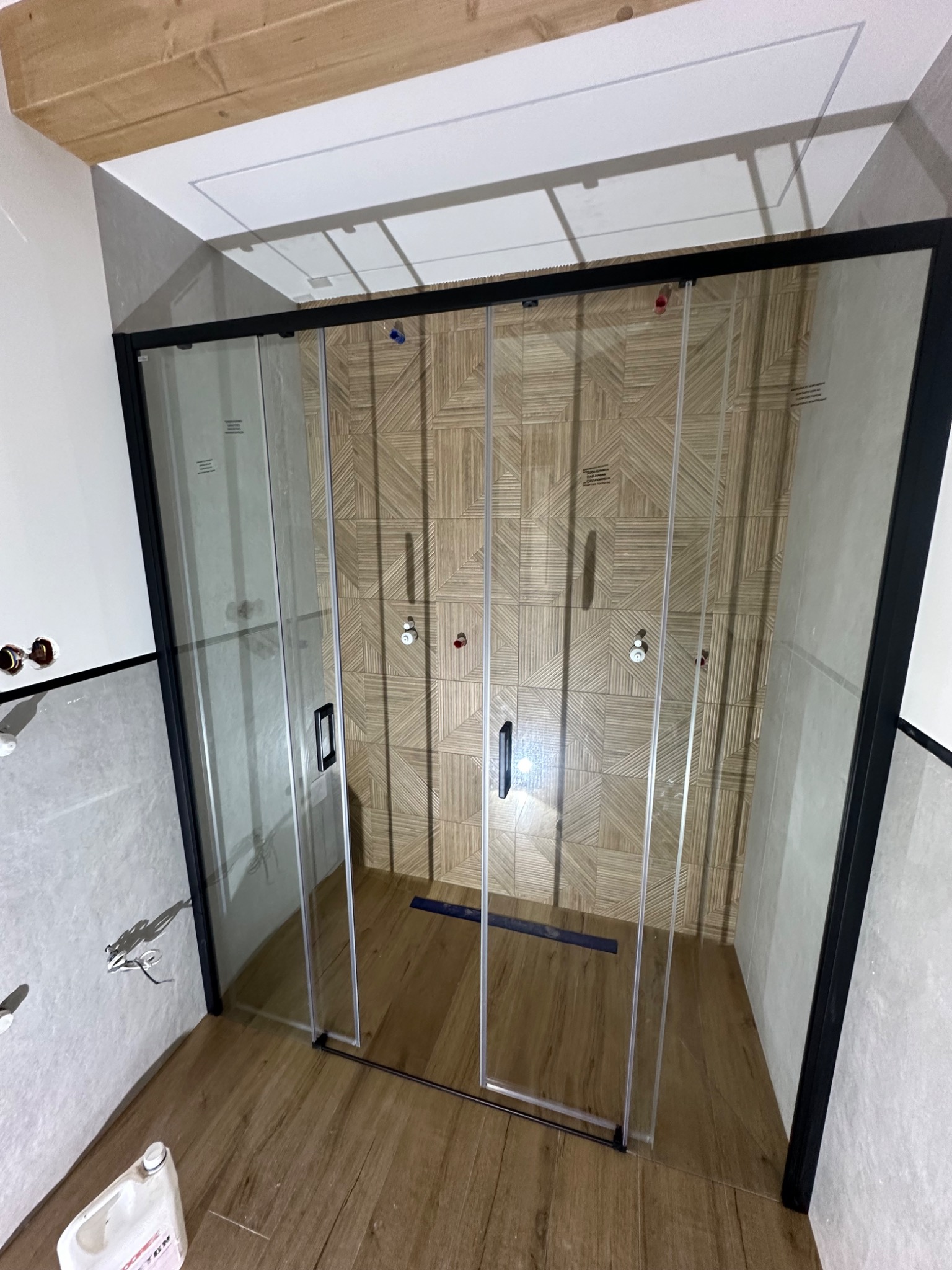 Nowoczesna kabina prysznicowa z czarnymi profilami i przesuwnymi drzwiami na tle ściany z dekoracyjną, drewnopodobną mozaiką. Widoczna butelka z detergentem na podłodze.