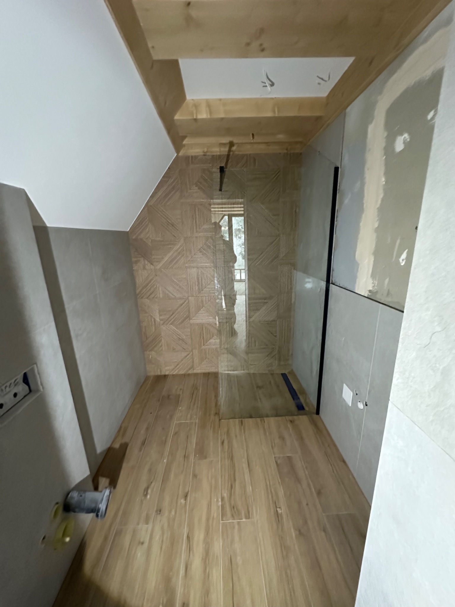 Wąska łazienka w trakcie wykańczania, z drewnopodobnymi panelami na podłodze i ścianie, częściowo wykończona glazurą, widoczna kabina prysznicowa z czarnymi profilami i drewniana konstrukcja sufitu.