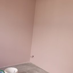 Fachowy Pracownik Budowlany - Różowe ściany świeżo po malowaniu, widoczne puszki farby Knauf Super na szarym, surowym betonie podłogi i wystający kabel elektryczny z sufitu w rogu pokoju.