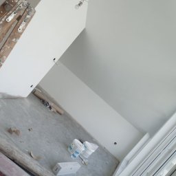 Fachowy Pracownik Budowlany - Wnętrze w trakcie odświeżania: pomalowane na biało ściany, sufit ze skosem, drabina malarska, puszki z farbą i surowa żarówka na kablu.