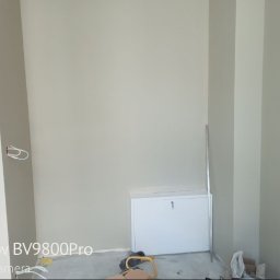 Fachowy Pracownik Budowlany - Świeżo pomalowane ściany na szaro, widoczne kable wystające ze ściany, biała szafka pod ścianą i fragment łazienki w głębi