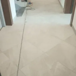 Jasne, kwadratowe płytki podłogowe ułożone diagonalnie w korytarzu, widoczne przewody elektryczne i przedłużacz, fragment schodów z czarną balustradą w tle.