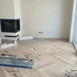 Ułożona podłoga z paneli w jodełkę, widoczna przecinarka do paneli, w tle kominek z czarnymi elementami i surowe ściany.