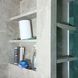 Fachowy Pracownik Budowlany - Półki wnękowe w łazience wyłożone płytkami imitującymi marmur, widoczne akcesoria montażowe i środki czystości, zielone płytki w tle.