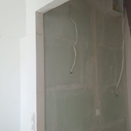 Fachowy Pracownik Budowlany - Ściana z płyt gipsowo-kartonowych w trakcie szpachlowania, widoczne wystające kable elektryczne i fragment instalacji wodnej, surowy stan wnętrza.