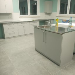 Fachowy Pracownik Budowlany - Jasna kuchnia z szarymi szafkami, metalowym blatem i chromowaną baterią, widok na okno i płytki podłogowe, butelka wody i płyn do naczyń na blacie.