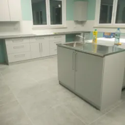 Jasna kuchnia z szarymi szafkami, metalowym blatem i chromowaną baterią, widok na okno i płytki podłogowe, butelka wody i płyn do naczyń na blacie.