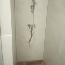 Kabina prysznicowa wyłożona jasnymi, kwadratowymi płytkami, z brązowymi płytkami imitującymi drewno na podłodze i odpływem liniowym. Widoczna bateria prysznicowa z deszczownicą i słuchawką.