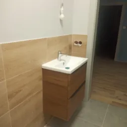 Nowoczesna szafka łazienkowa z umywalką nablatową, drewnopodobne płytki na ścianie, niedokończone podłączenie elektryczne żarówki, szara podłoga i fragment drewnianej podłogi w przejściu do innego...