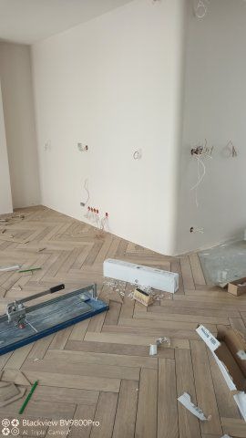 Ułożona drewniana podłoga w jodełkę w jasnym pokoju, z widocznymi puszkami elektrycznymi w ścianach i narzędziem do cięcia płytek na podłodze.