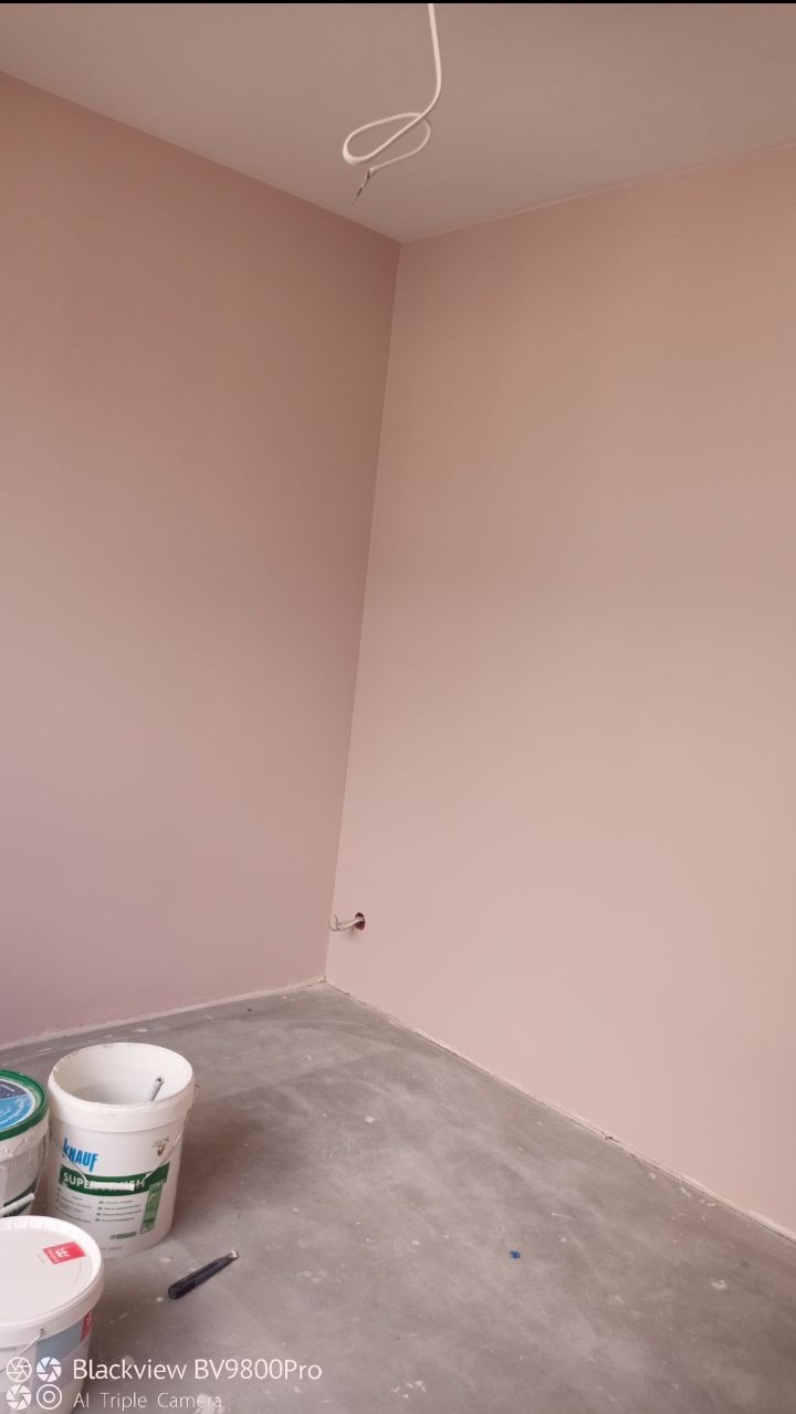 Różowe ściany świeżo po malowaniu, widoczne puszki farby Knauf Super na szarym, surowym betonie podłogi i wystający kabel elektryczny z sufitu w rogu pokoju.