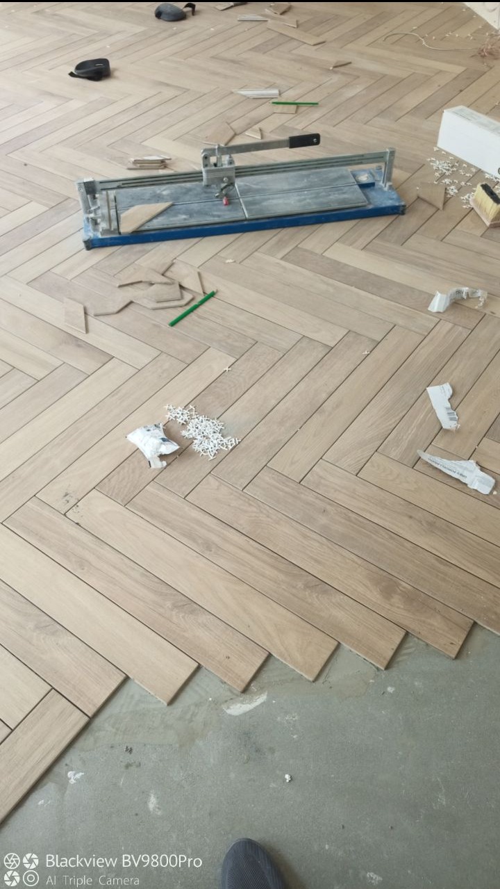 Układanie jasnych paneli podłogowych w jodełkę, widoczna przecinarka do paneli, ołówek i pozostałości po cięciu.