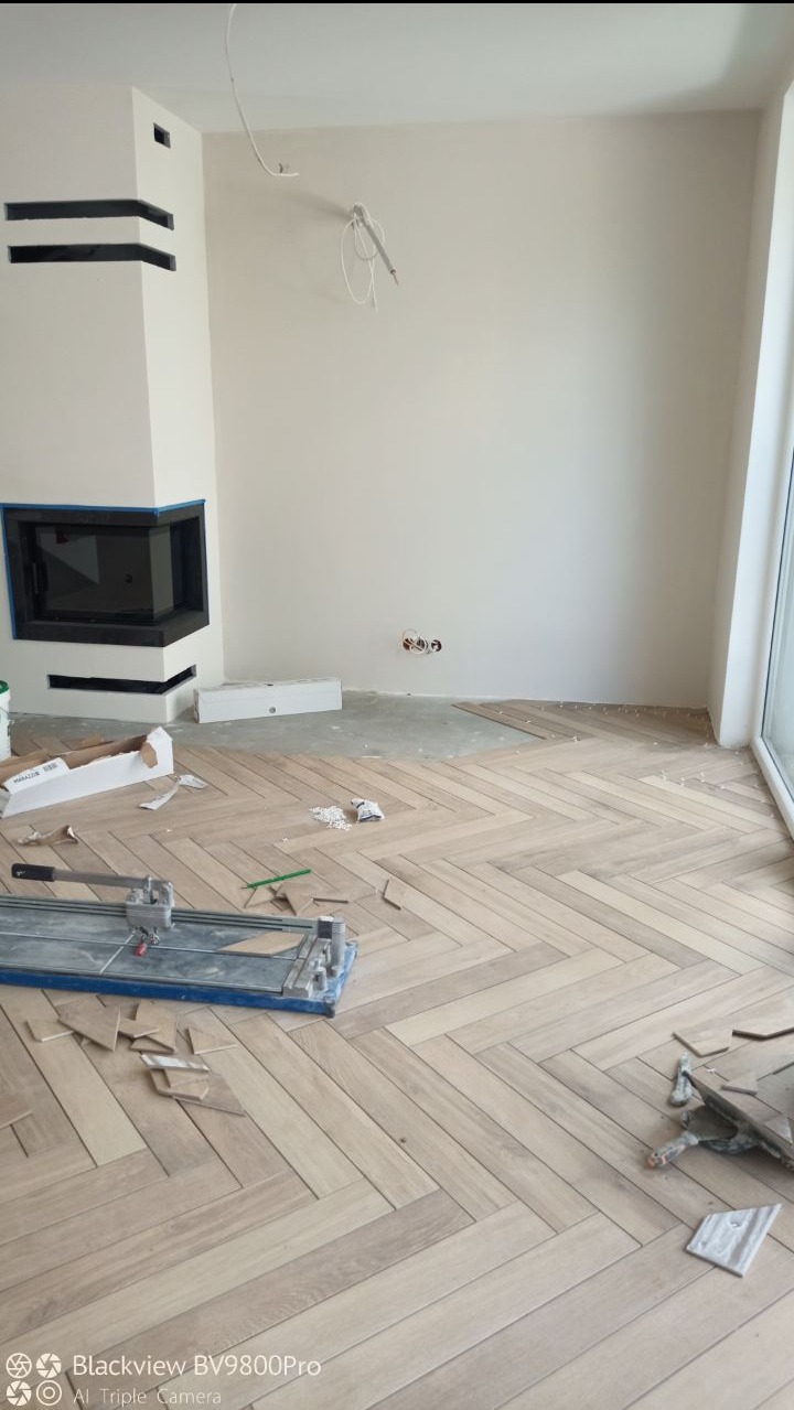 Układanie parkietu w jodełkę w jasnym pomieszczeniu z kominkiem, widoczna przecinarka do płytek i resztki materiału.