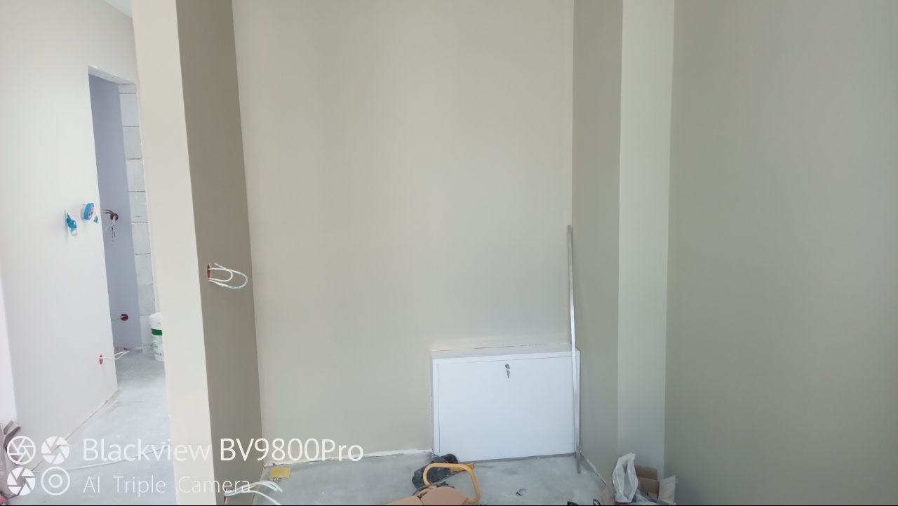 Świeżo pomalowane ściany na szaro, widoczne kable wystające ze ściany, biała szafka pod ścianą i fragment łazienki w głębi