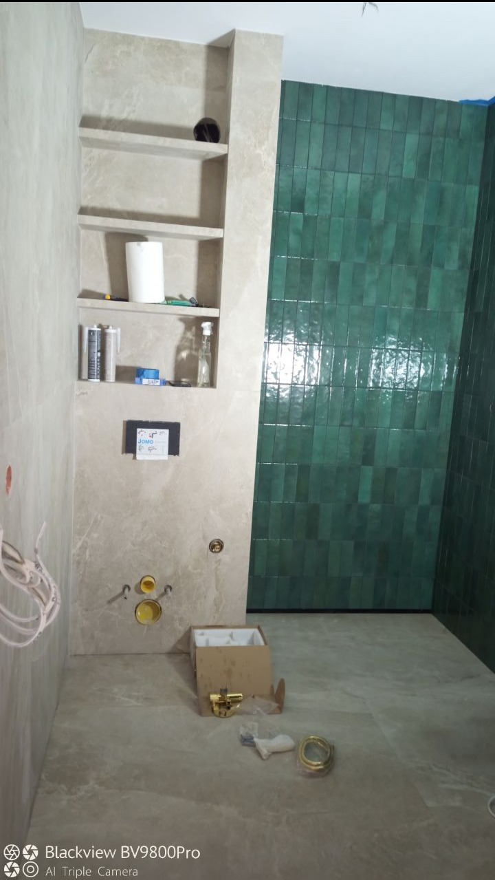 Wnętrze łazienki w trakcie prac wykończeniowych: zielona, pionowo ułożona glazura na ścianie, wnęka z półkami z beżowego marmuru, elementy instalacji sanitarnej w kartonie na podłodze.