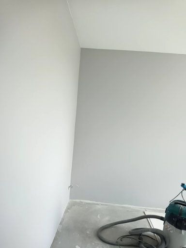 Świeżo pomalowane ściany na szaro w rogu pokoju, widoczny odkurzacz przemysłowy i kabel przy podłodze.