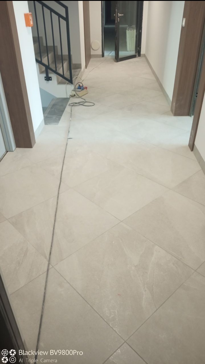 Jasne, kwadratowe płytki podłogowe ułożone diagonalnie w korytarzu, widoczne przewody elektryczne i przedłużacz, fragment schodów z czarną balustradą w tle.