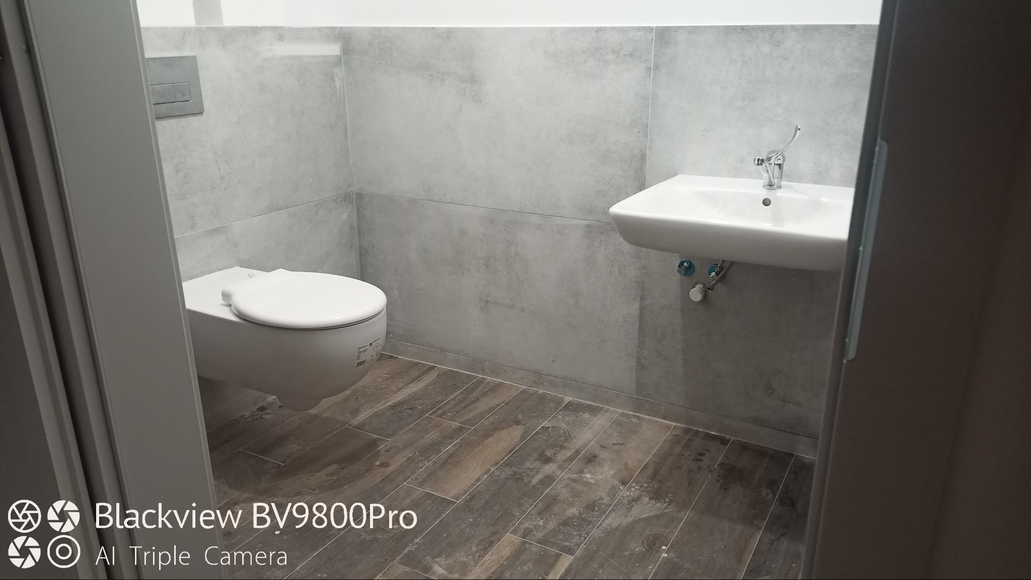 Nowoczesna łazienka z szarymi płytkami imitującymi beton, podwieszana toaleta i umywalka z chromowaną baterią, drewnopodobna podłoga z płytek.