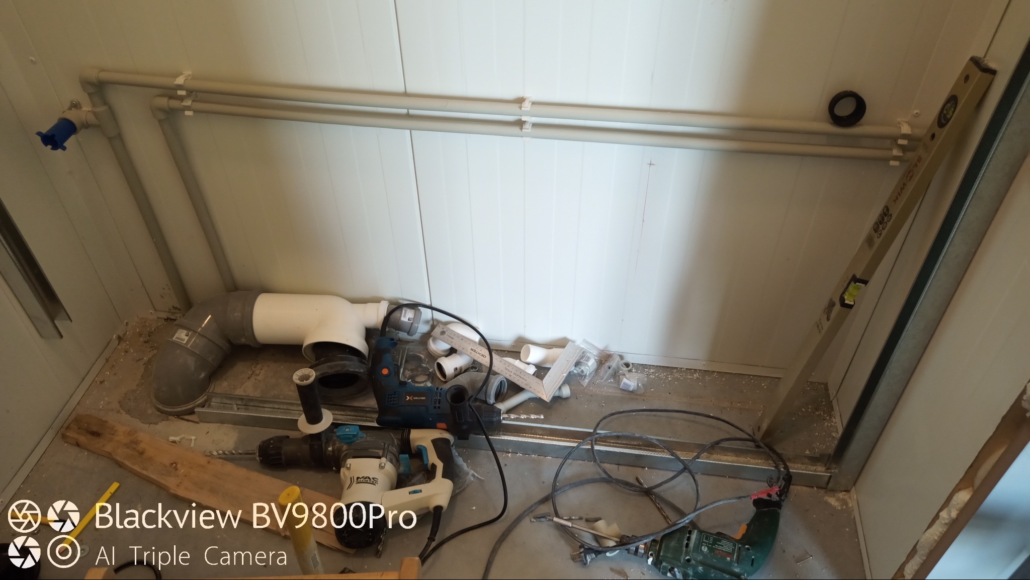 Wnętrze pomieszczenia w trakcie prac instalacyjnych: widoczne rury PCV, poziomica oparta o ścianę, wiertarki, przewody elektryczne i akcesoria instalacyjne na podłodze.