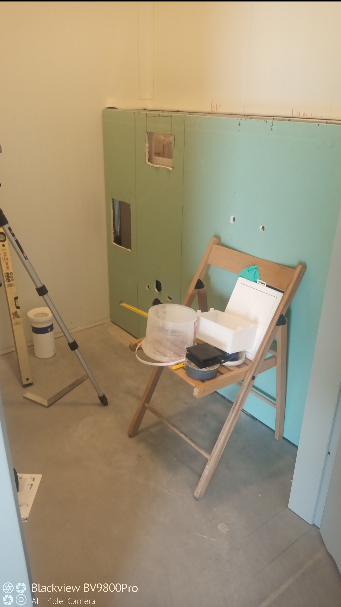 Ściana z płyt kartonowo-gipsowych z wyciętymi otworami, poziomica oparta o ścianę, krzesło z narzędziami i pojemnikami malarskimi.