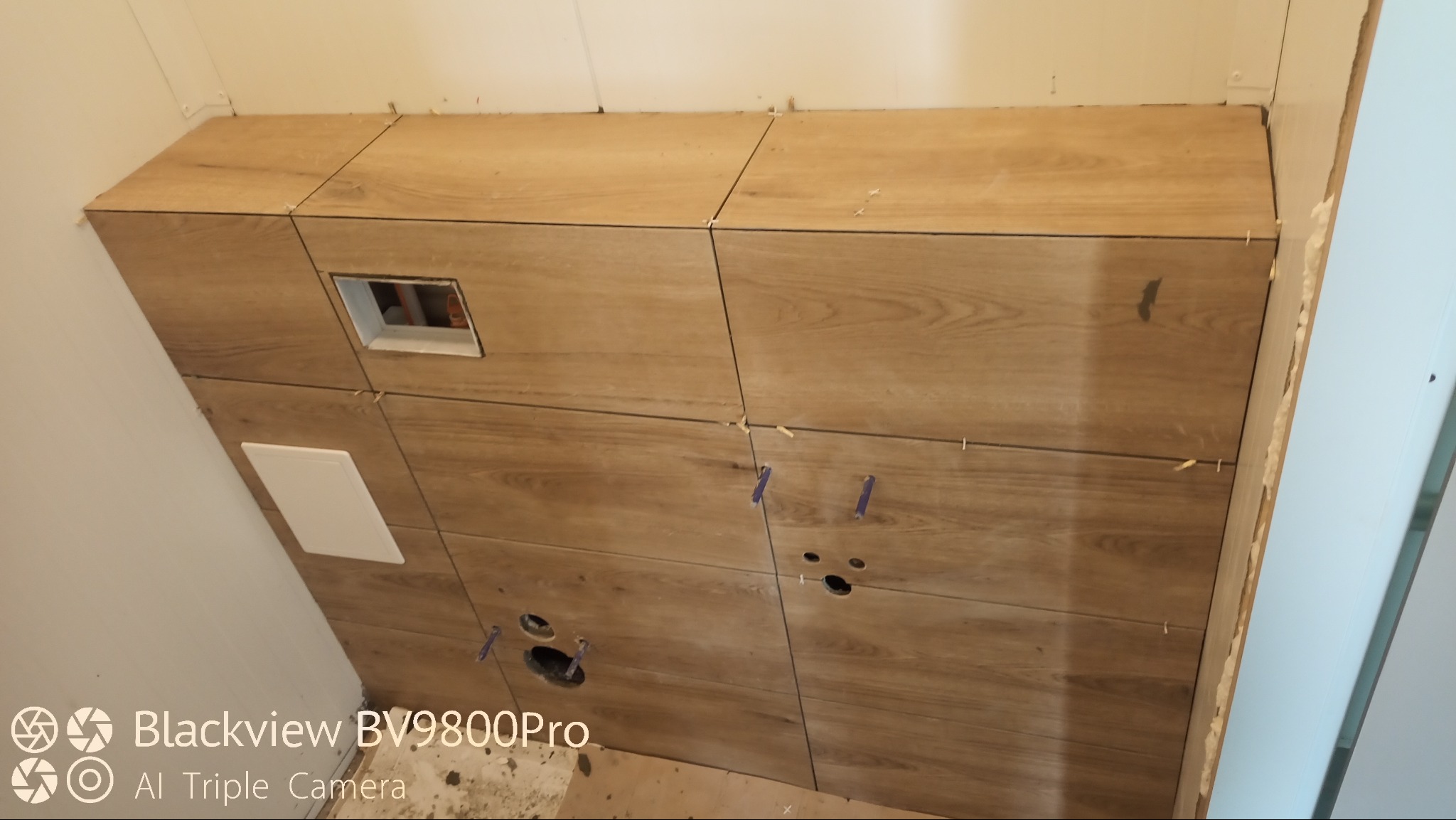 Obudowa stelaża WC wykończona płytkami imitującymi drewno, widoczne otwory na instalacje i biały otwór rewizyjny, fugi zabezpieczone krzyżykami dystansowymi.