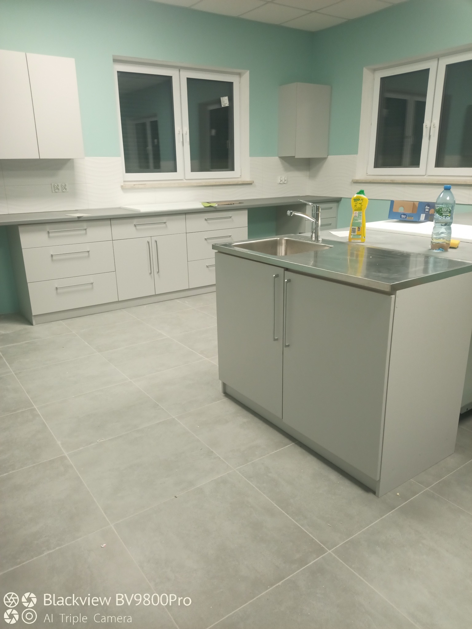 Jasna kuchnia z szarymi szafkami, metalowym blatem i chromowaną baterią, widok na okno i płytki podłogowe, butelka wody i płyn do naczyń na blacie.