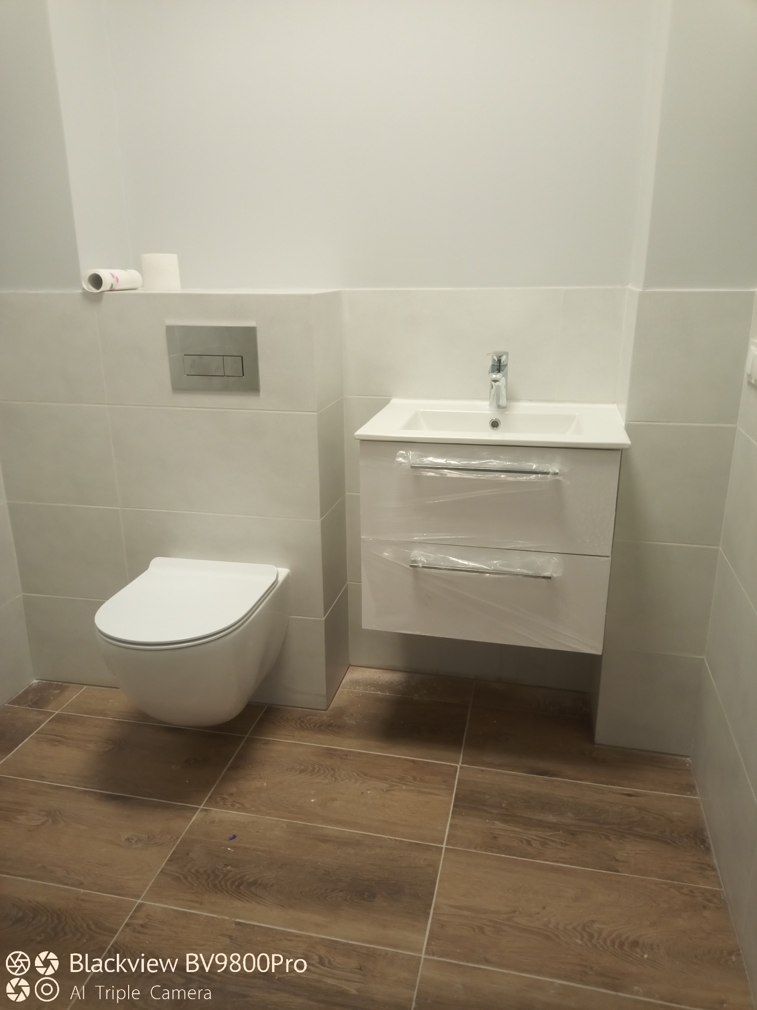 Nowoczesna łazienka z wiszącą toaletą, szafką z umywalką w folii ochronnej i brązowymi płytkami imitującymi drewno na podłodze.