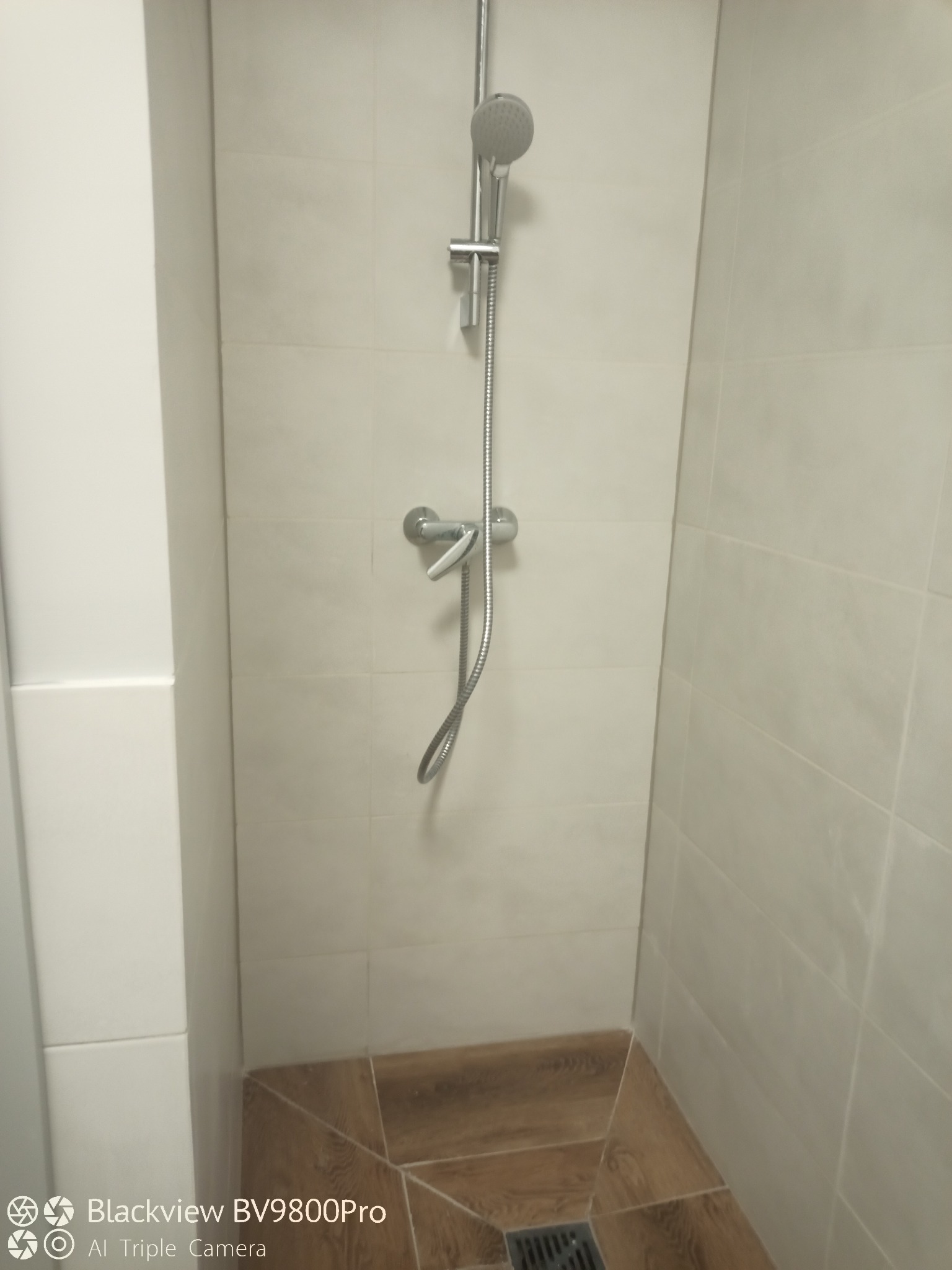 Kabina prysznicowa wyłożona jasnymi, kwadratowymi płytkami, z brązowymi płytkami imitującymi drewno na podłodze i odpływem liniowym. Widoczna bateria prysznicowa z deszczownicą i słuchawką.