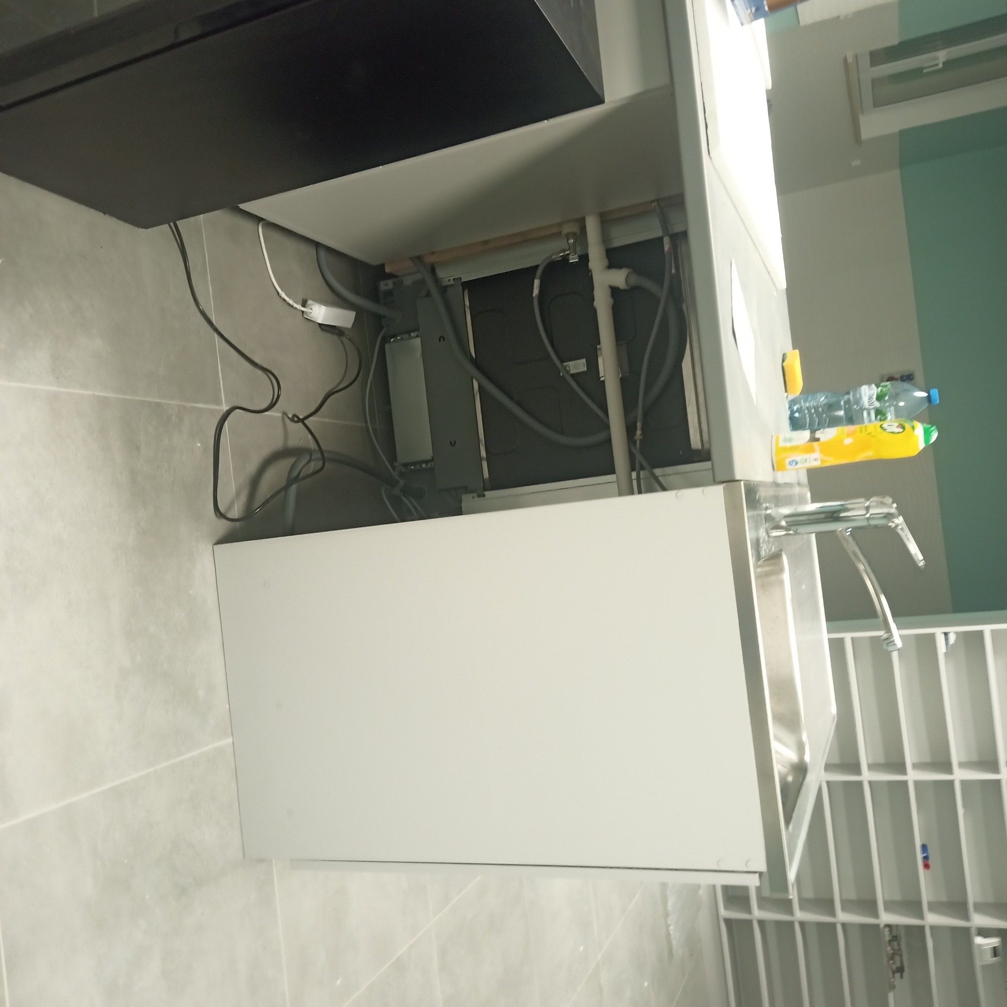 Nowoczesna szafka pod umywalkę z widocznymi instalacjami hydraulicznymi i elektrycznymi, butelki z płynami obok baterii, białe płytki na podłodze i ścianie.