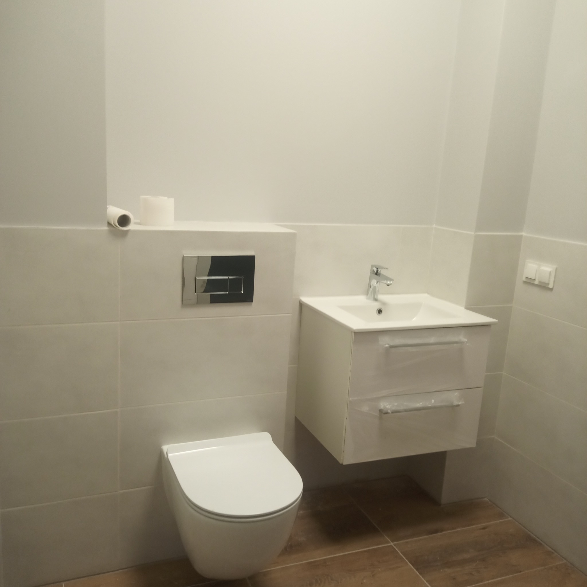 Nowoczesna łazienka z podwieszaną toaletą, umywalką z szafką i brązowymi płytkami podłogowymi, widoczne zabezpieczenia folią na szafce.