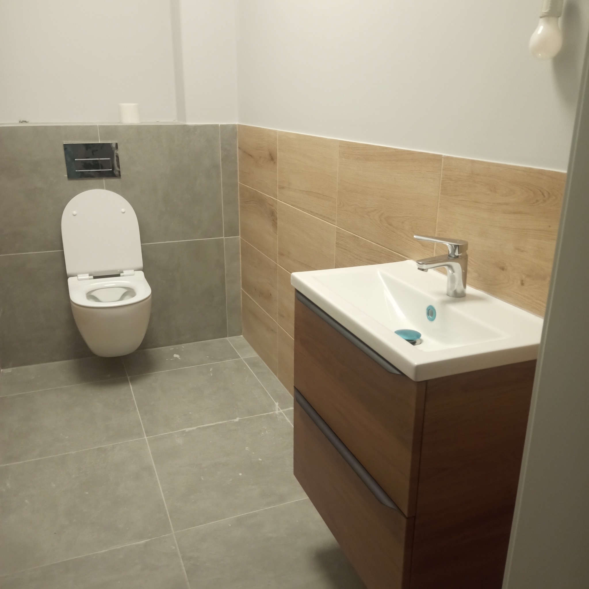 Nowoczesna łazienka z podwieszaną toaletą, szarą płytką podłogową i drewnopodobną okładziną ścienną za umywalką z szafką.