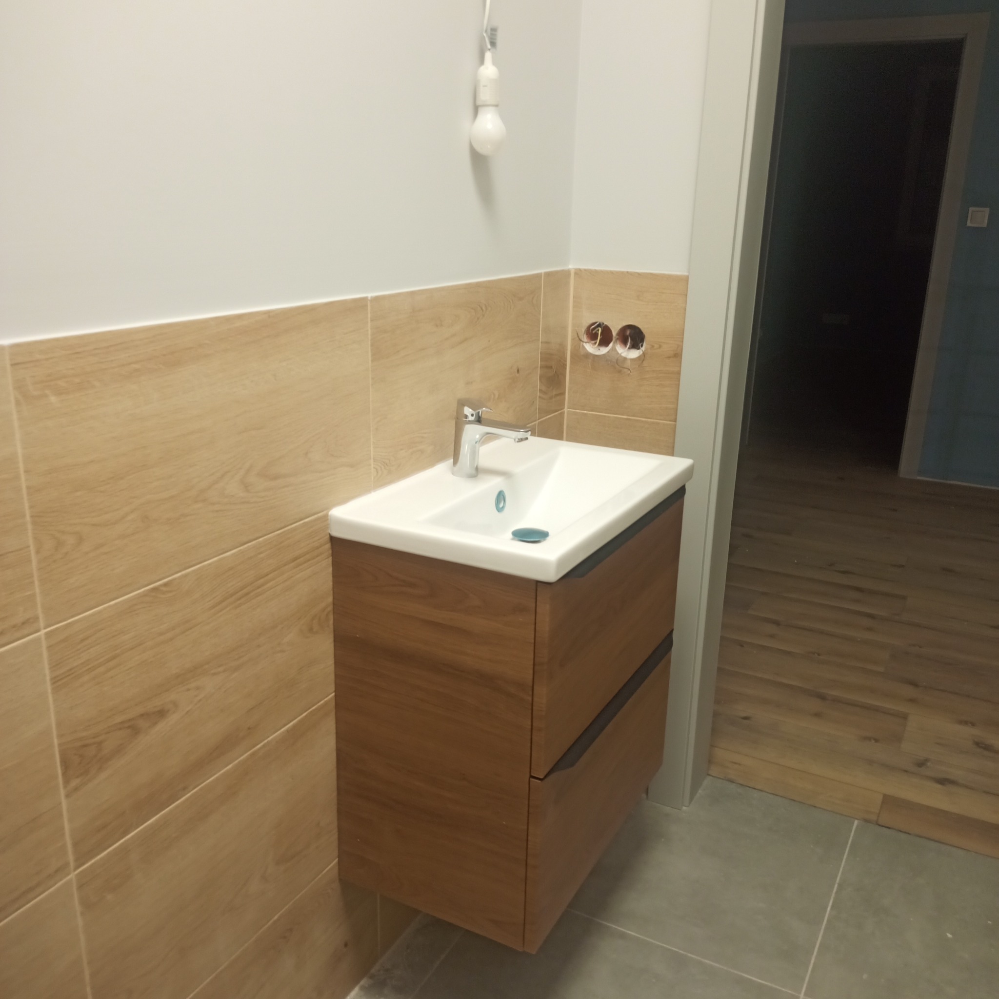 Nowoczesna szafka łazienkowa z umywalką nablatową, drewnopodobne płytki na ścianie, niedokończone podłączenie elektryczne żarówki, szara podłoga i fragment drewnianej podłogi w przejściu do innego...