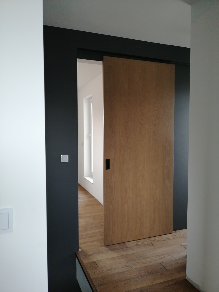 Dębowa płyta przesuwna w systemie bez widocznej szyny jezdnej, zamontowana w ciemnej futrynie, widoczna z perspektywy wnętrza, z podłogą wykończoną w drewnie.