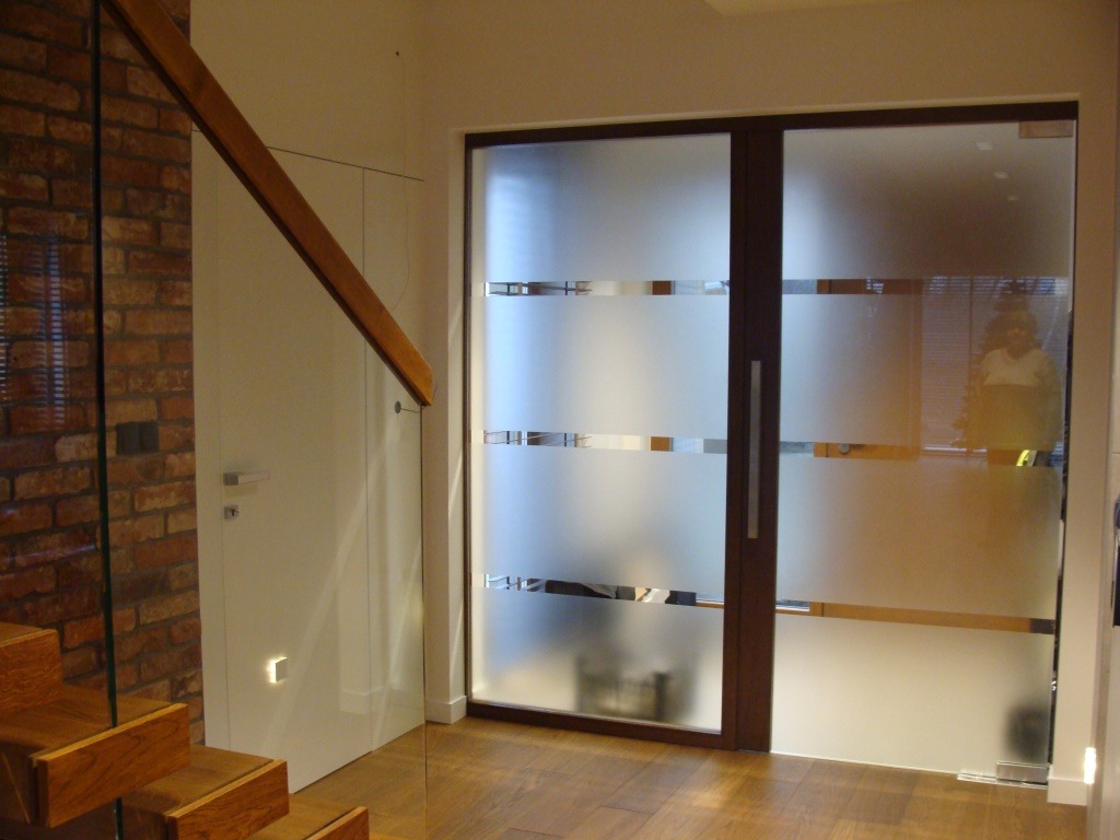 Szklane drzwi przesuwne z poziomymi paskami dekoracyjnymi, oddzielające korytarz od salonu, widoczne drewniane schody i ceglana ściana w tle.
