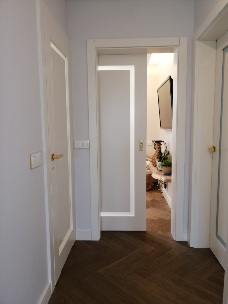 Białe drzwi wewnętrzne przesuwne z delikatną ramką, widok z perspektywy wejścia na parkiet ułożony w jodełkę, w tle rośliny doniczkowe i telewizor.