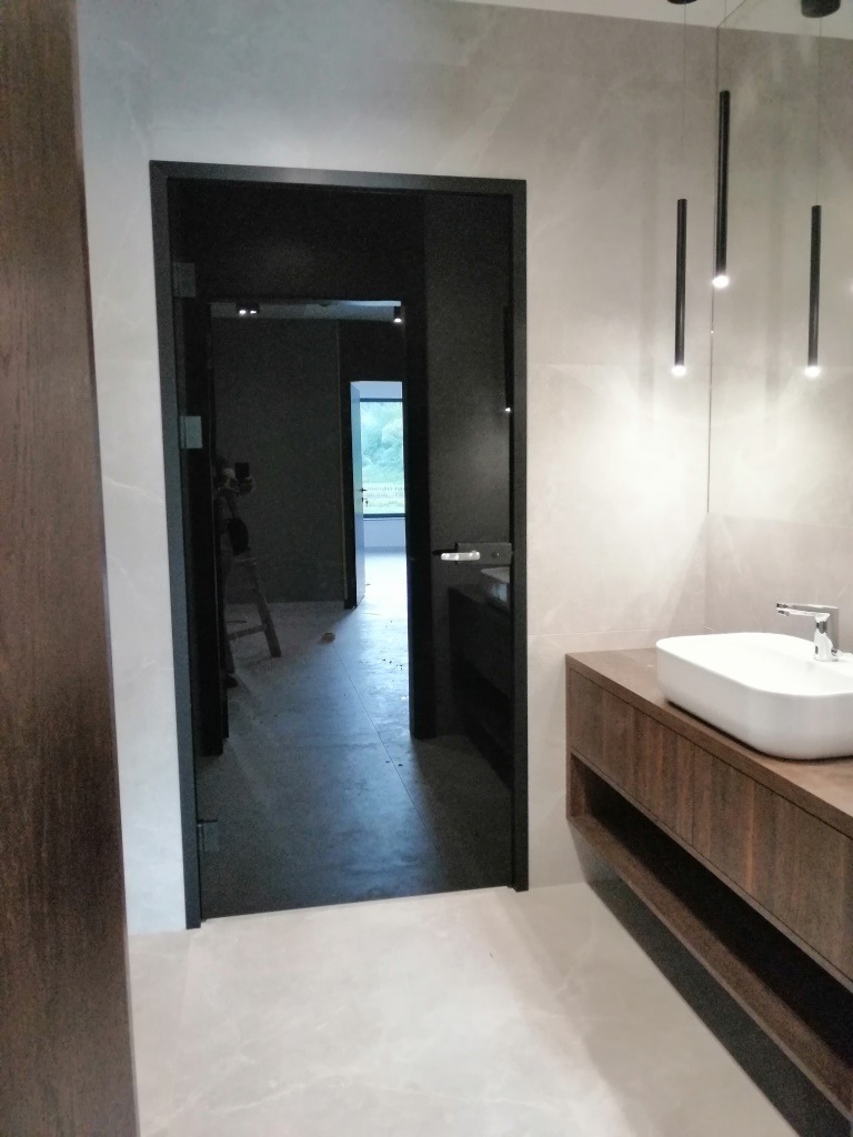 Czarne, szklane skrzydło w łazience z umywalką na drewnianej szafce i minimalistycznymi lampami wiszącymi.