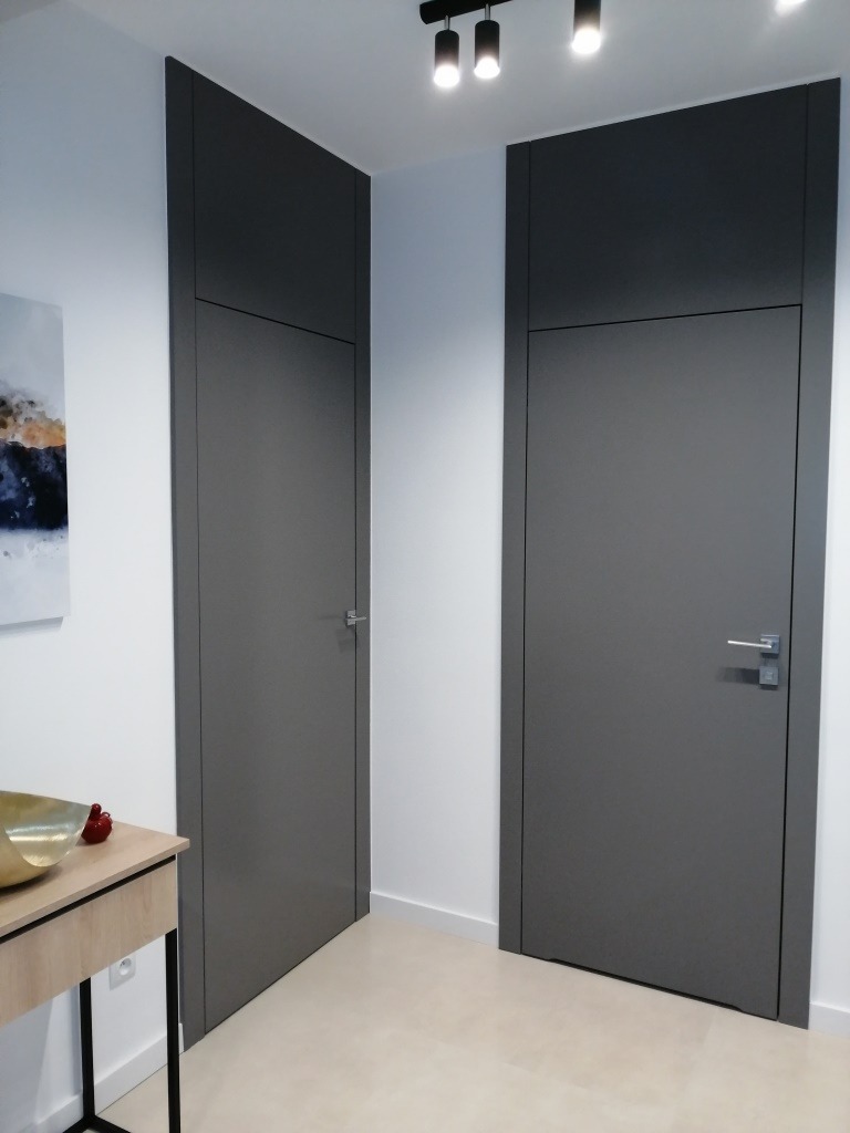 Dwa wysokie, popielate skrzydła drzwiowe, sięgające do sufitu z nadstawką, umieszczone w białej ścianie, widoczne klamki i oświetlenie sufitowe.