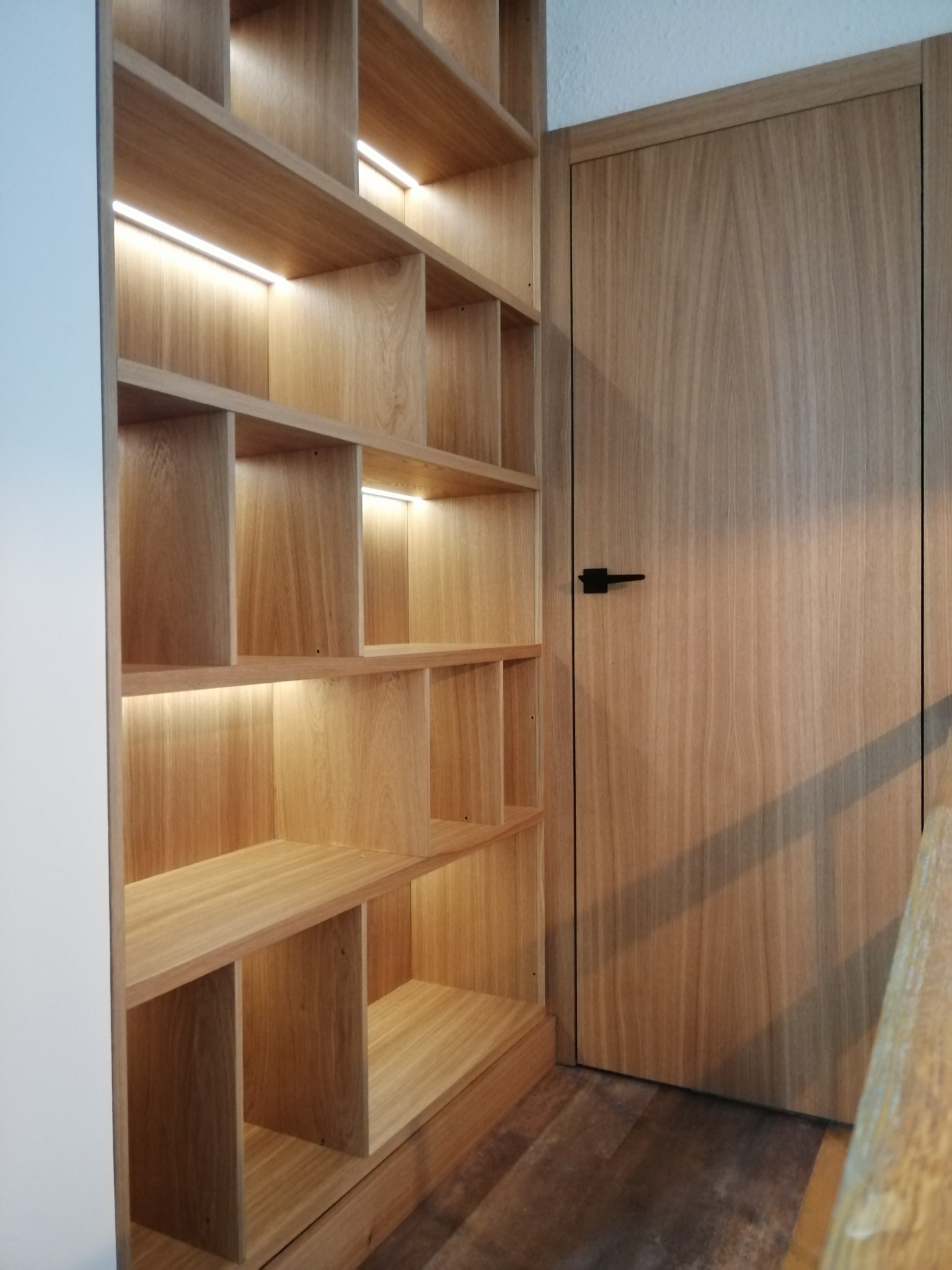 Dębowa zabudowa biblioteki z podświetlanymi półkami, zintegrowana z dębowymi drzwiami z czarną klamką.